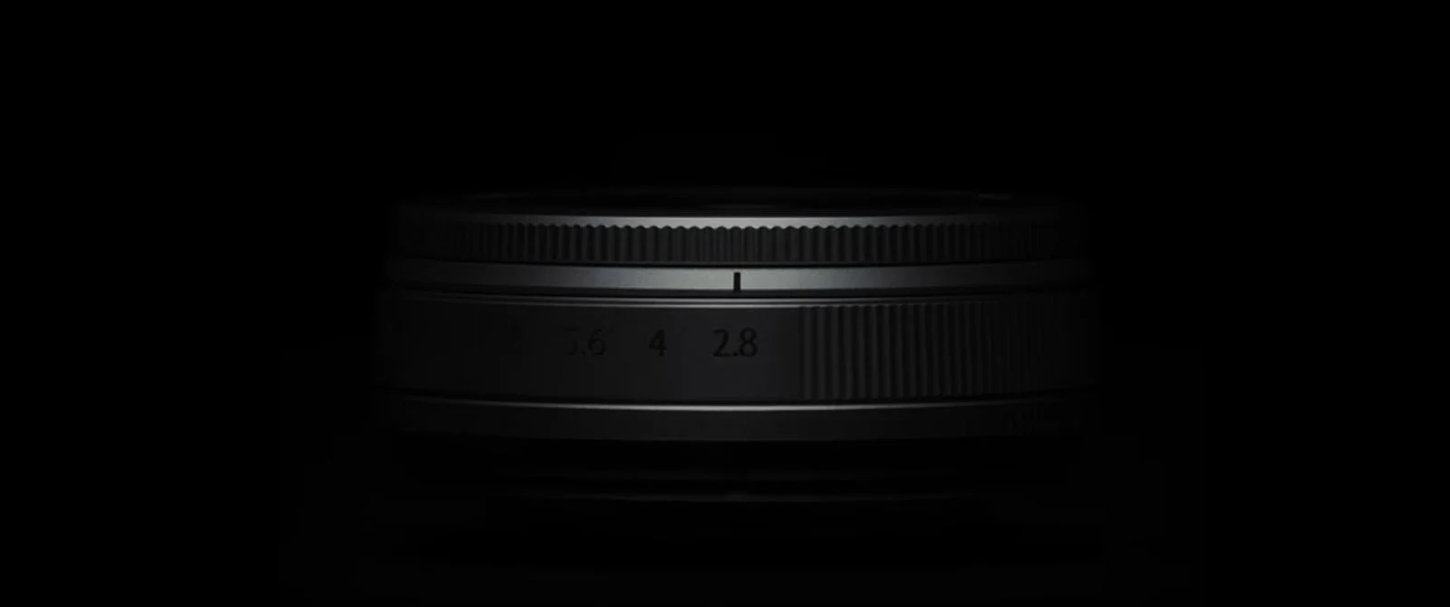 a camera lens is partially illuminated against a black background with its ridged focus and zoom rings visible and aperture mar